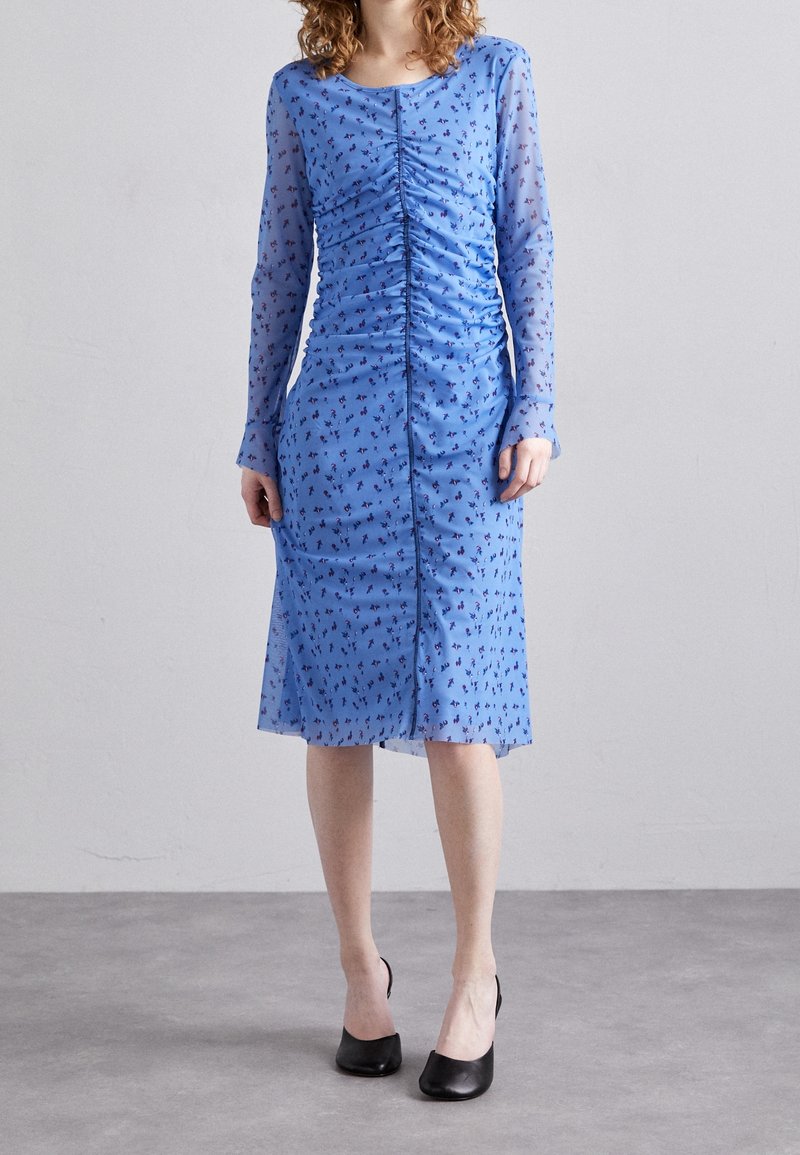 Femme portant une robe bleue au genou avec des manches longues en voile, un petit imprimé floral, un devant froncé et des chaussures noires à talons bas.