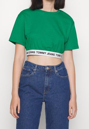 Kobieta ubrana w zielony krótki t-shirt z "Tommy Jeans" na dole i niebieskie dżinsy z wysokim stanem na jednolitym tle.