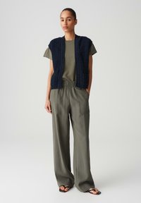 Gilet lavorato a maglia blu marina sopra una maglietta verde oliva, abbinato a pantaloni larghi di un leggero colore oliva. L'outfit presenta una vestibilità rilassata e un tessuto testurizzato.