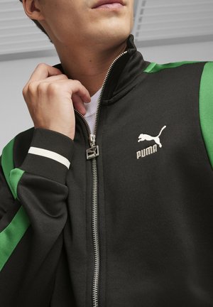 Giacca nera con accenti verdi, colletto alto, zip completa e logo Puma. Polsi a coste e dettaglio a strisce bianche sulle maniche.