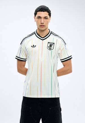 Fiatal férfi fehér focimezben, melyen többszínű függőleges csíkok, a japán labdarúgó címer és fekete Adidas logó látható, hátrafont kézzel áll.