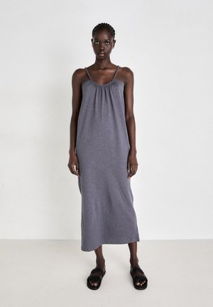 Grå, ærmeløs maxi-kjole med elastisk hals og rynket stoff, laget av lett materiale. Brukt med svarte, stroppede sandaler.