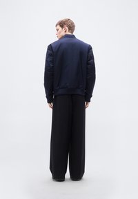 Σκούρο μπλε μπουφάν bomber με ριμπ γιακά και μανσέτες, με καπιτονέ ώμους, συνδυασμένο με φαρδιά μαύρη παντελόνα. Απλός, καθαρός σχεδιασμός και υφή.