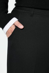 Černé kalhoty s hladkou texturou a boční kapsou, doplněné černým žebrovaným svetrem s bílým manžetem.