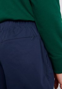Navyblauwe broek met een gestructureerd oppervlak, elastische tailleband en een discreet ritszakje aan de rechterkant. Groene trui zichtbaar aan de bovenkant.