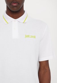 Balta polo marškiniai iš tekstūruoto audinio, su žaliai geltonais apykaklės akcentais ir išsiuvinėtu "Justcavalli" logotipu kairėje krūtinės pusėje.