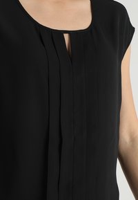Blusa nera con scollatura rotonda, dettaglio plissettato sul davanti e maniche corte. Realizzata in tessuto liscio con una leggera lucentezza.
