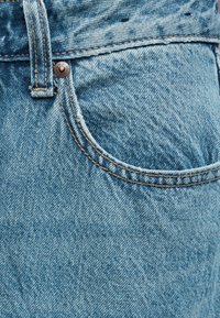 Κοντινή λήψη ανοιχτό μπλε τζιν, που δείχνει μπροστινή τσέπη, θηλιά για ζώνη, λεπτομέρειες ραφών και μεταλλικό κουμπί με ορειχάλκινο καρφί.