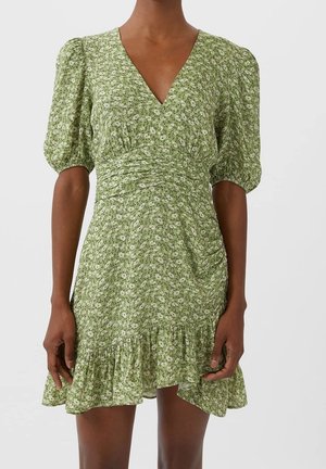 Femme portant une robe verte à fleurs avec col en V, manches bouffantes, taille froncée et ourlet à volants sur fond uni.