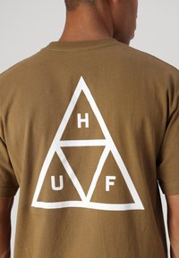 Camiseta de algodón marrón con un gran gráfico triangular blanco en la parte posterior, que presenta las letras H, U y F dentro de las formas.