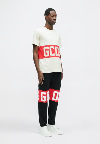 T-shirt grigio chiaro con una striscia orizzontale rossa che presenta "GCDS" in grande lettere bianche; abbinato a pantaloni della tuta neri con dettagli rossi.