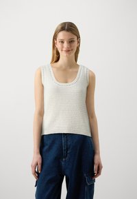Valge kudumine tank top, millel on ümar kaelus ja soonikäär. Omab tekstuurset mustrit. Kantud lahtiste siniste teksapükstega.