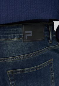 Σκούρα μπλε τζιν τζιν με ορατή πίσω τσέπη και μαύρη ορθογώνια ετικέτα με στυλιζαρισμένο λογότυπο "F", φορεμένα με μπλε υφασμάτινο πουλόβερ.