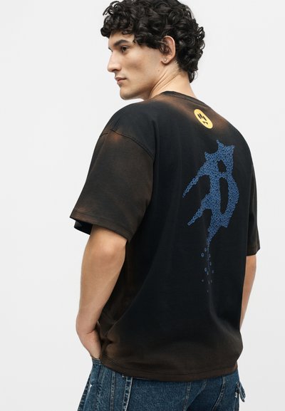 Άνδρας με κατσαρά μαλλιά που φοράει μαύρο υπερμεγέθες t-shirt με μπλε γραφικό σε στυλ φυσαλίδας και κίτρινο κυκλικό σύμβολο στην πλάτη, συνδυασμένο με μπλε τζιν.