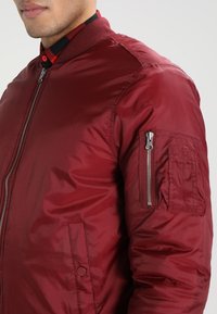 Burgundy bomberjacka med en slät, glänsande finish, ribbat krage och sidoficka. Utrustad med en dragkedjeförsedd armficka för förvaring.