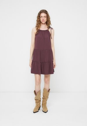 Young woman with wavy hair wearing sleeveless purple tiered dress and slouchy tan suede boots, standing against white background.