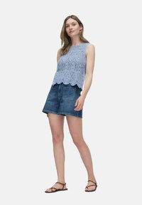 Ljusblå spetsblus med vågig nederkant, parat med mörka denimshorts. Modellen bär bruna remsandaler. Enkel vit bakgrund.