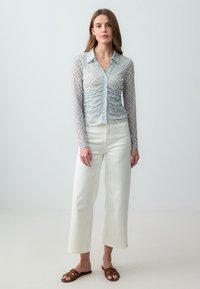 Blusa a scacchi azzurro chiaro con maniche lunghe trasparenti, chiusura a bottoni e dettagli arricciati, abbinata a pantaloni a gamba larga di colore bianco sporco e sandali marroni.