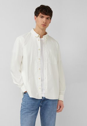 Junger Mann, der ein aufgeknöpftes weißes Langarmhemd über einem weißen T-Shirt und blauen Jeans trägt, vor einem schlichten grauen Hintergrund stehend.