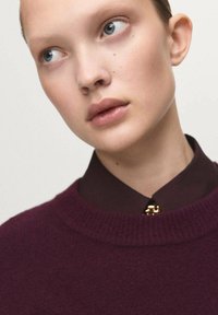 Maglione borgogna a maglia con collo a coste indossato sopra una camicia marrone con colletto, caratterizzato da un dettaglio con bottone dorato. Texture liscia e design aderente.