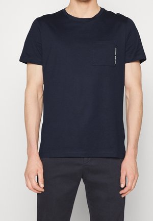 Homme portant un T-shirt à manches courtes bleu marine uni avec une poche poitrine et un petit texte blanc vertical sur le bord de la poche.