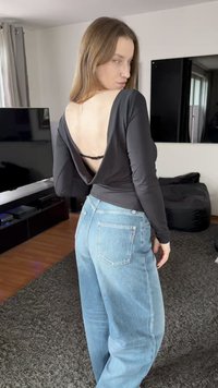 Femme aux cheveux longs portant un haut noir à dos nu et un jean bleu clair, debout dans un salon avec une télévision et un pouf.