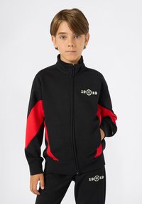Ragazzo che indossa una giacca nera con zip e dettagli rossi e pantaloni abbinati, entrambi con il logo "19 19", in posa con una mano in tasca.