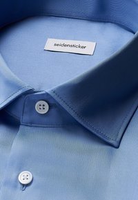 Camicia azzurra chiara con colletto classico, leggera lucentezza e tre bottoni bianchi. Etichetta del marchio "seidensticker" visibile vicino al colletto.