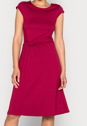 Kobieta w bezrękawnej sukience sięgającej do kolan w kolorze magenta z marszczonym detalem w talii, stojąca na jednolitym jasnym tle.
