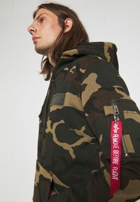 Sötétzöld és bézs camo kapucnis felső, pamut keverékből készült. Zsebében cipzáras ujjal és piros "Remove Before Flight" címkével rendelkezik.