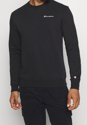 Mann trägt schwarzen Champion-Sweatshirt mit Logo auf der Brust und am Ärmel, kombiniert mit schwarzer Hose, steht vor einfarbigem Hintergrund.