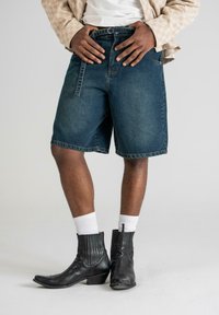 Shorts en denim bleu foncé avec une coupe droite et des détails de couture visibles, associés à des bottines noires et des chaussettes blanches côtelées.