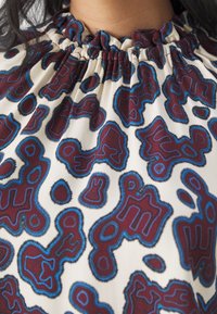 Tessuto crema con scollo arricciato presenta motivi astratti in bordeaux e blu, offrendo un design texturizzato e colorato.