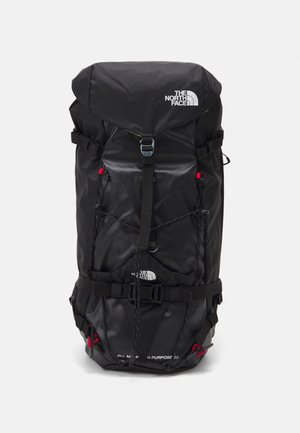 Mochila de nylon negra con solapa superior ajustable, múltiples correas y acentos rojos. Presenta un logotipo y varios compartimentos para almacenamiento.