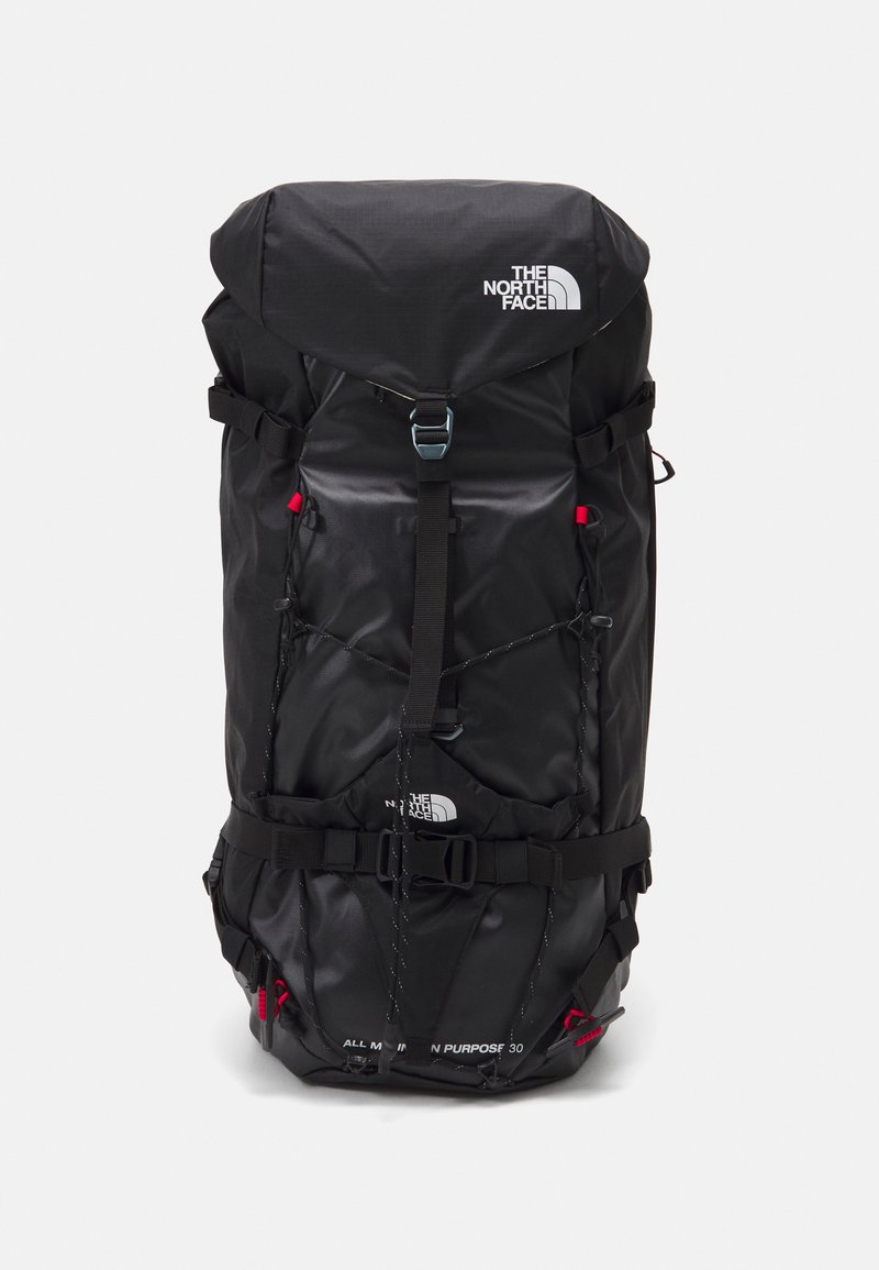 Mochila de nylon negra con solapa superior ajustable, múltiples correas y acentos rojos. Presenta un logotipo y varios compartimentos para almacenamiento.