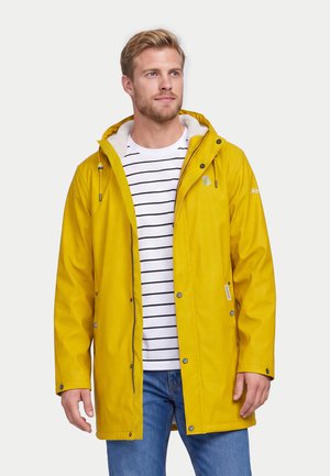 Man draagt een gele regenjas over een wit-zwart gestreept shirt met blauwe spijkerbroek, staand tegen een effen achtergrond.