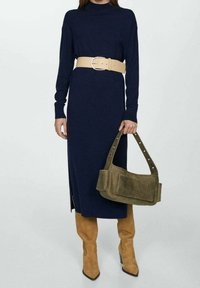 Vestido de manga larga azul marino con un cuello alto, ceñido con un cinturón ancho beige. Combinado con botas beige hasta la rodilla y un bolso verde de hombro.