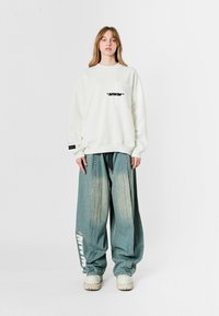 Sudadera blanca de gran tamaño con un bolsillo en el pecho; presenta texto impreso en negro. Pantalones de mezclilla sueltos en azul claro con un logo blanco en el costado y cordones.