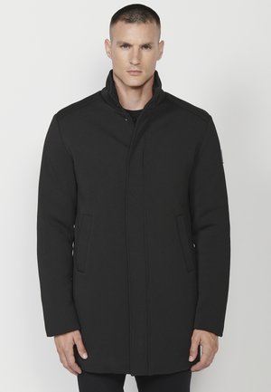 Schwarzer Mantel mit hohem Kragen, Knopfverschluss und seitlichen Taschen. Aus strukturiertem Stoff mit klaren Linien und minimaler Detailgestaltung.