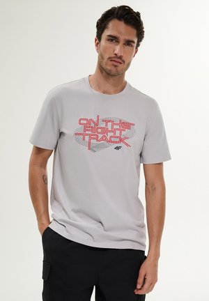 Mężczyzna stoi, ubrany w jasnoszary T-shirt z czerwonym napisem "ON THE RIGHT TRACK" i czarne spodnie, neutralny wyraz twarzy, jednolite tło.
