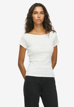 Femme aux longs cheveux bouclés portant un haut blanc à manches courtes et un pantalon noir, debout devant un fond blanc uni.