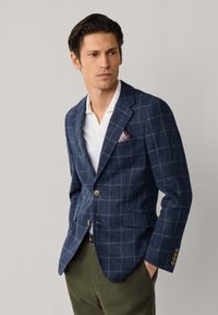 Muž v modrom kockovanom saku s bielou košeľou a olivovo zelenými nohavicami stojaci na jednoduchom svetlom pozadí.