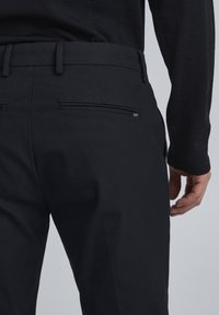 Svarta byxor med en slät textur, utrustade med en sidoficka och en logotagg. Midjebandet är åtsittande och ger en skräddarsydd look.