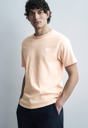 Νέος άνδρας με σγουρά μαλλιά που φοράει ανοιχτό ροδακινί T-shirt με μικρό λογότυπο καρδιάς στο στήθος και σκούρο παντελόνι, στέκεται με το ένα χέρι στην τσέπη.