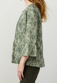 Grön blus med mönster och blommönster, tre fjärdedelsärmar och en avslappnad passform. Har en rund halsringning och plisserade detaljer.