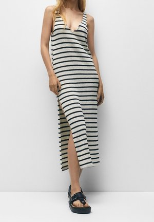 Femme portant une robe tricotée sans manches à rayures blanches et noires avec une fente latérale, associée à des sandales compensées noires.