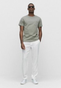 Kortärmad, olivgrön t-shirt med bröstficka, tillsammans med vita linnbyxor och vita sneakers. Modellen bär också svarta solglasögon.