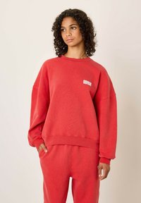 Sarkans džemperis ar apaļu kakla izgriezumu, lielgabarīta piegriezums, ribota piedurkne un apakšmala, ar mazu baltu logo priekšpusē. Mīksta, teksturēta auduma.