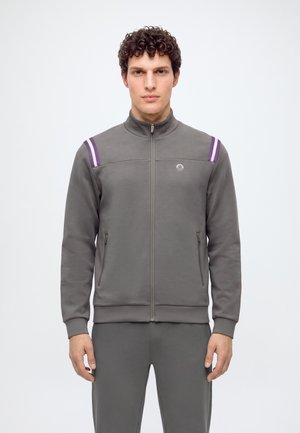 Homme aux cheveux bouclés portant une veste grise zippée avec des rayures violettes et blanches sur les épaules et un pantalon gris assorti sur fond blanc.