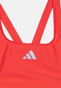 Crvena atletska sportska brassiere sa uskim trakama i bijelim adidas logotipom sprijeda. Glatki, rastezljivi materijal s bešavnim dizajnom.